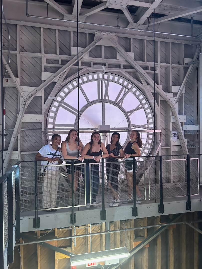 A group of five people are leaning against a railing. Behind them is the outline of a large clock. They are in the inside of a clock tower.