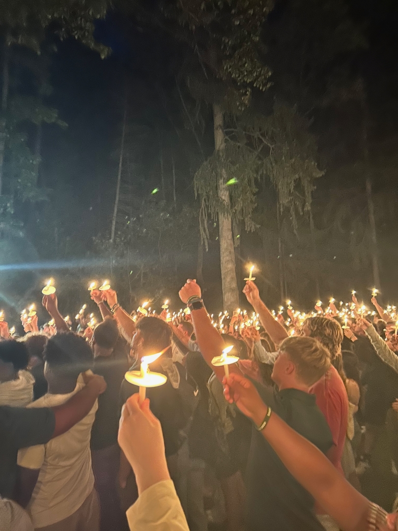 a group of people are gathered. They are holding up lit candles. There are large trees in the background