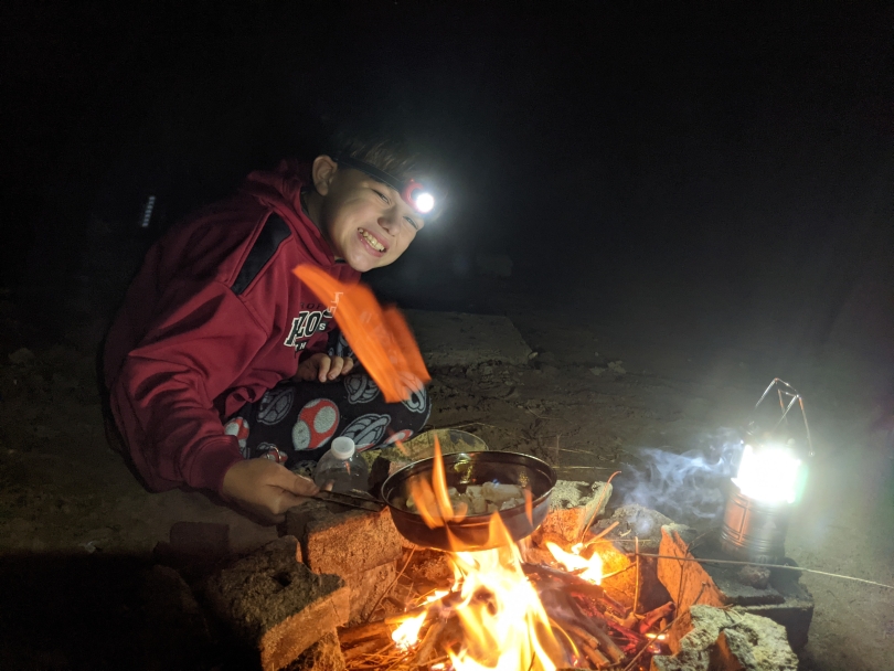 My brother around a campfire cooking some chicken while we camp by the riverbank