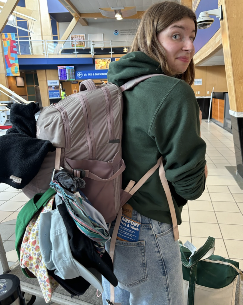 A girl in a green sweatshirt is looking at the camera. She is wearing a large backpack with many baseball caps attached to the outside of the bag.