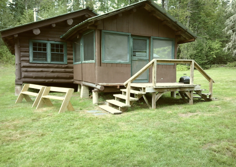 A brown two room cabin resting above a grassy field with trees in the back.
