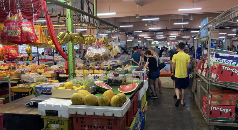 A view of a Malaysian fresh market, various fruits are displayed in a stand in the foreground. People are crowded around various stalls in the background.