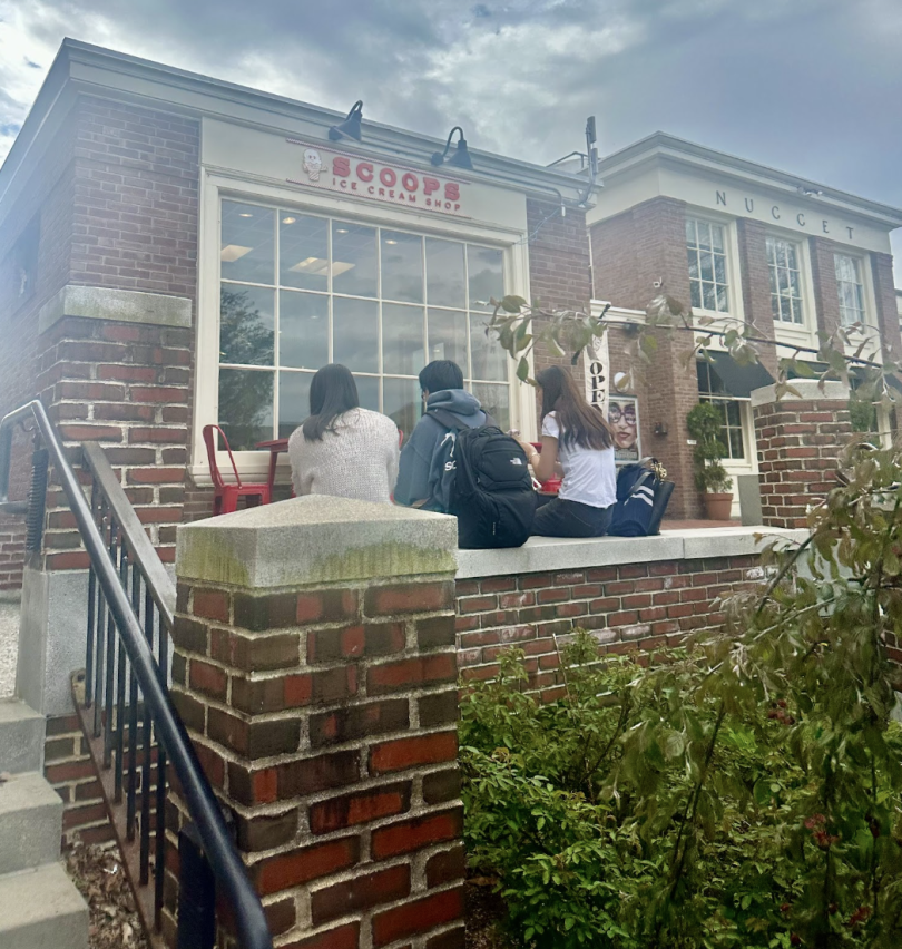 Three friends enjoying ice cream on a bench outside Scoops, a popular ice cream shop. The shop's colorful sign is visible in the background.