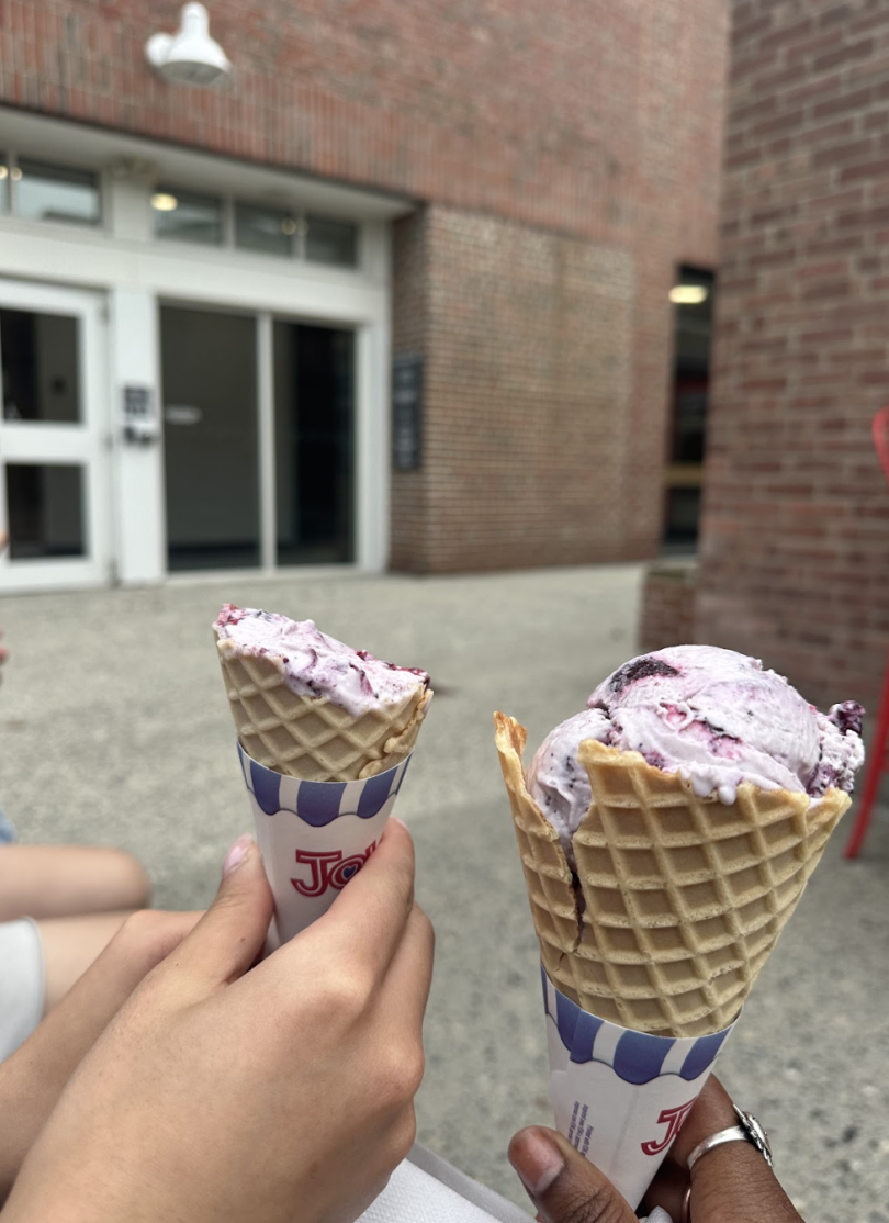 Two people holding up waffle cones filled with blueberry ice cream in front of the brick Scoops building. 