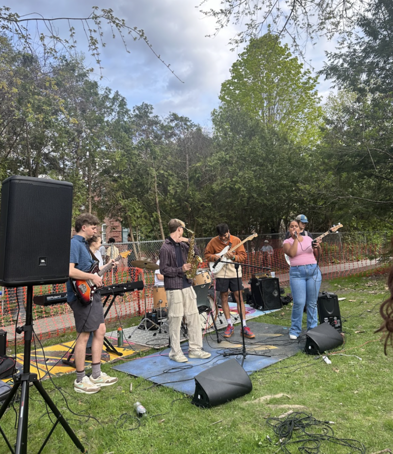 A student band performing on a grassy area at a Greek organization event. The band members are engaged in their performance.