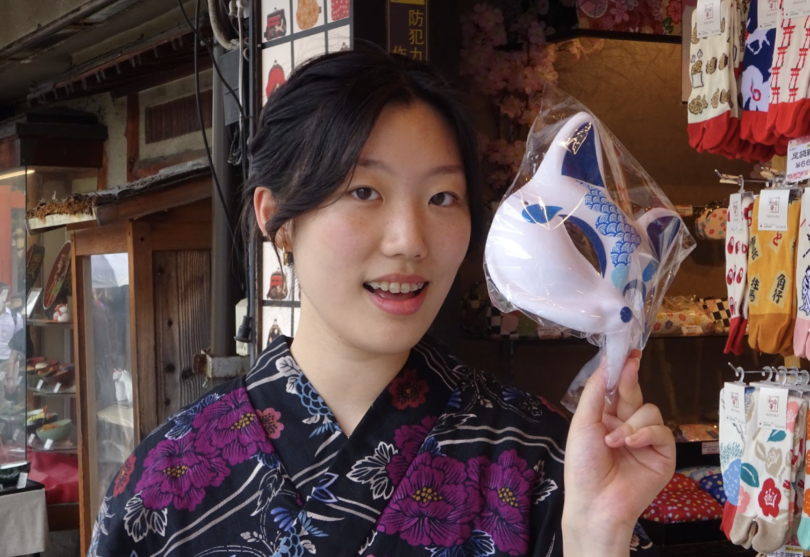 A portrait of a girl in traditional Japanese clothing smiling to the camera. She is holding a cartoon character mask.