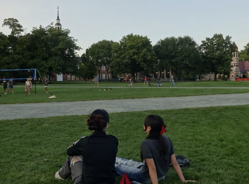 Two girls sit side-by-side on grass with their backs to the camera. There are people playing volleyball as well as a library in the background.