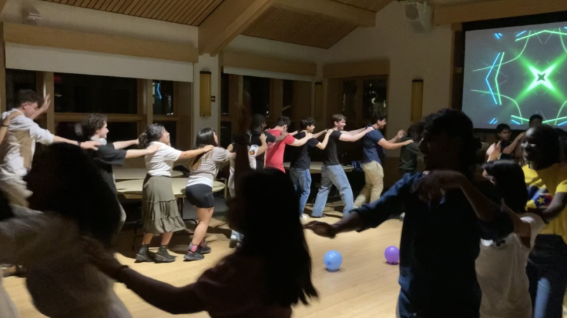 FYSEP students and staff participate in a conga line, wrapping around the picture. The foreground is dimly lit, while the background is bright with a blue and a purple balloon scattered on the floor. 