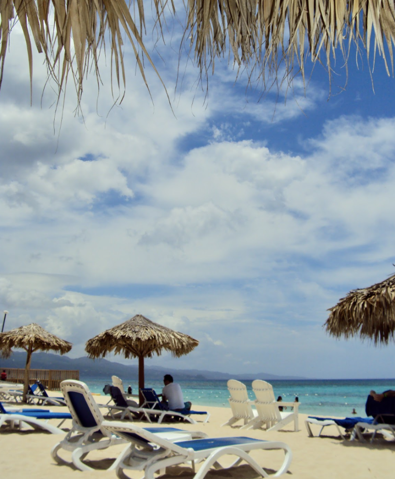 Beach with glistening blue water, sand dotted with beach chairs, and tiki umbrellas providing shade.