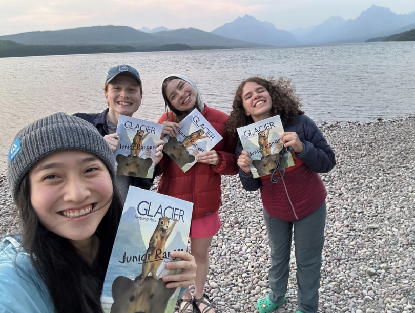 Four women smile for a selfie next to a lake. They are all holding up booklets that are Junior Ranger books for Glacier National Park.