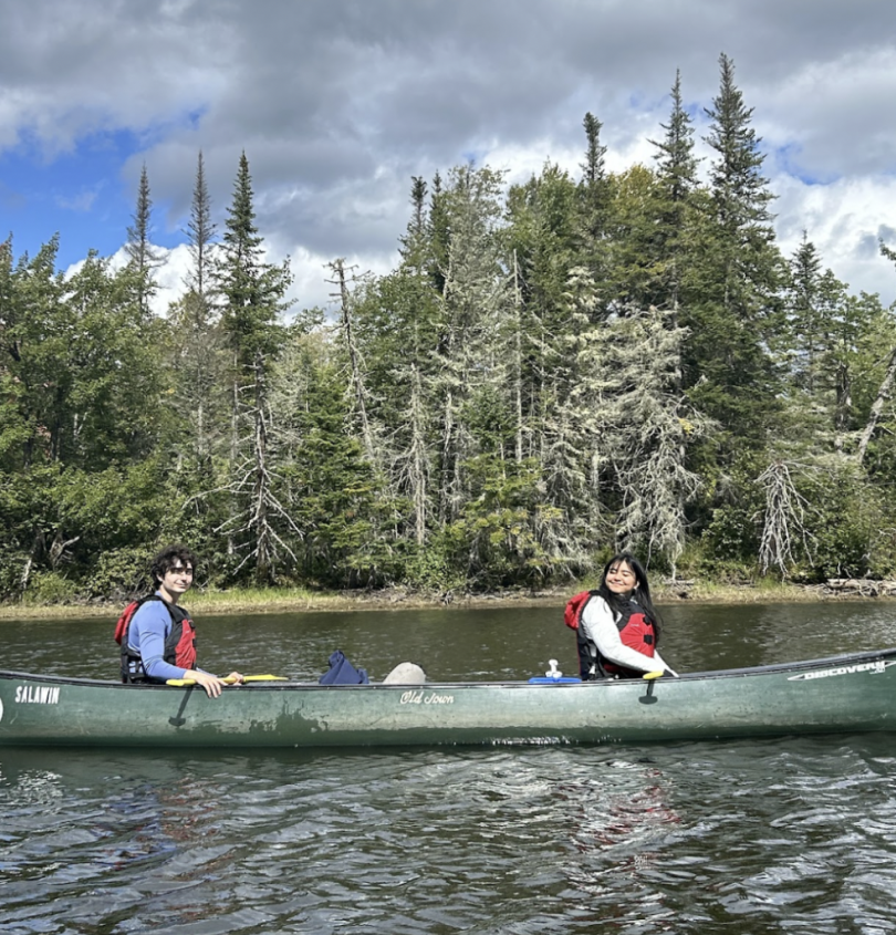 Two students in a canoe on a river, with evergreen trees along the riverbank behind them