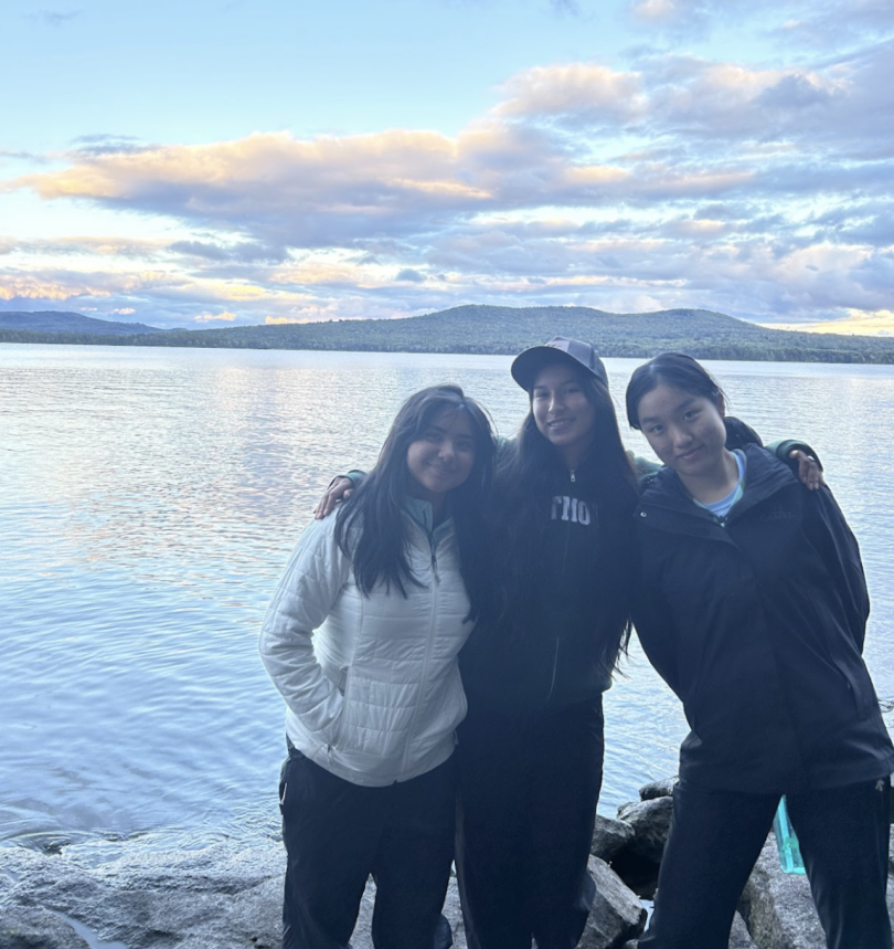 3 girls stand shoulder to shoulder on a rocky lakeshore during sunrise. Mountains, clouds, and a blue sky in the background.