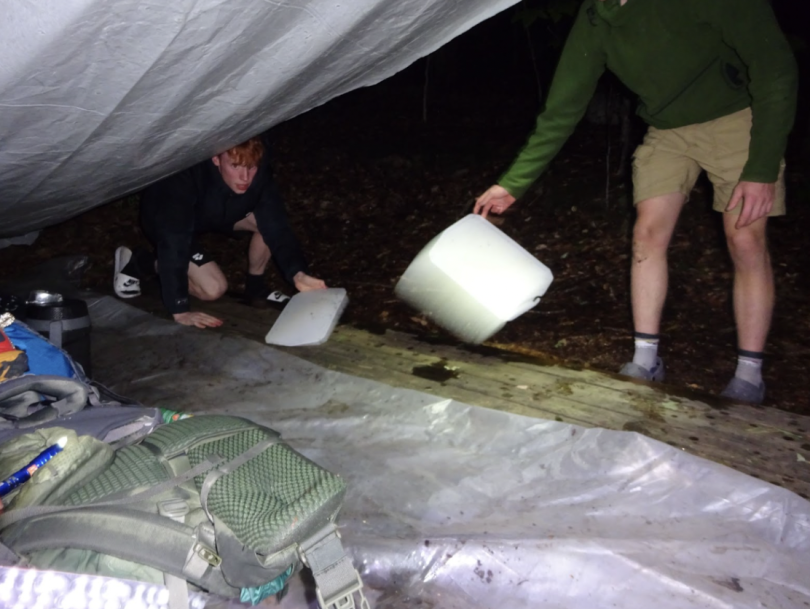 Inside a tent on a wooden platform in the dark, two people are working hard to scrape off the rain leakage in the tent. 