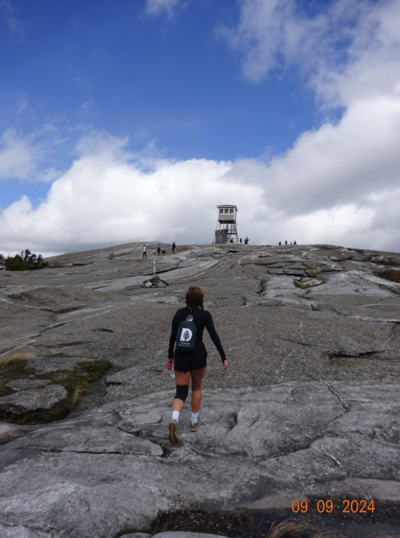 On the summit of a rocky mountain; a girl with her back to the camera hikes towards the tower in the distance.