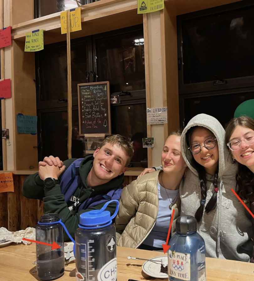 four people are sitting together at a table. They are smiling and have their arms around each other. Their water bottles are on the table in front of them along with their half-eaten plates of food.