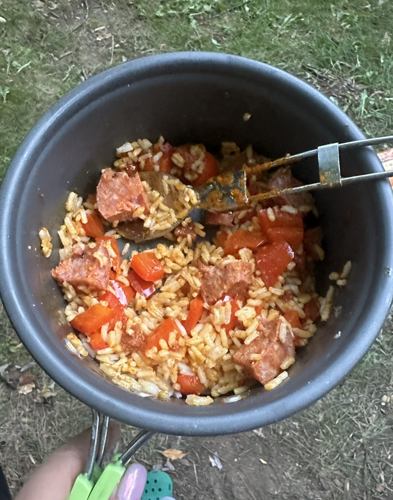 An image of me, Crystal, holding a camping pot containing rice, sausage, red bell peppers, and curry paste. There is also a spoon in the pot.