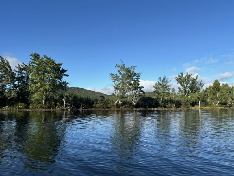 A picture of the Grant River with blue water and multiple trees and mountains in the background. The sky is blue, the sun is shining, and there are a few clouds in the background.