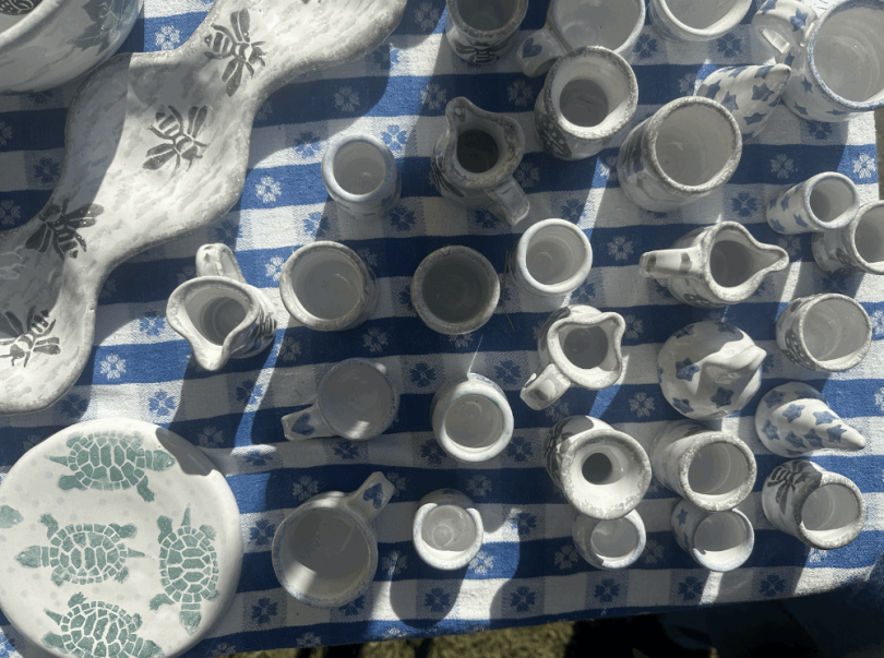 An aerial image of multiple sculpted pottery pieces including a plate with painted turtles, a dipping dish with painted bees, and numerous smaller pots and pitchers that have a painted design. The background is a blue and white striped tablecloth.