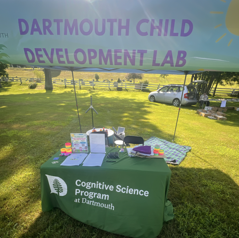 Lab booth under a tent with a sign reading "Dartmouth Child Development Lab." A desk is set up with a tablecloth displaying "Cognitive Science Program at Dartmouth."