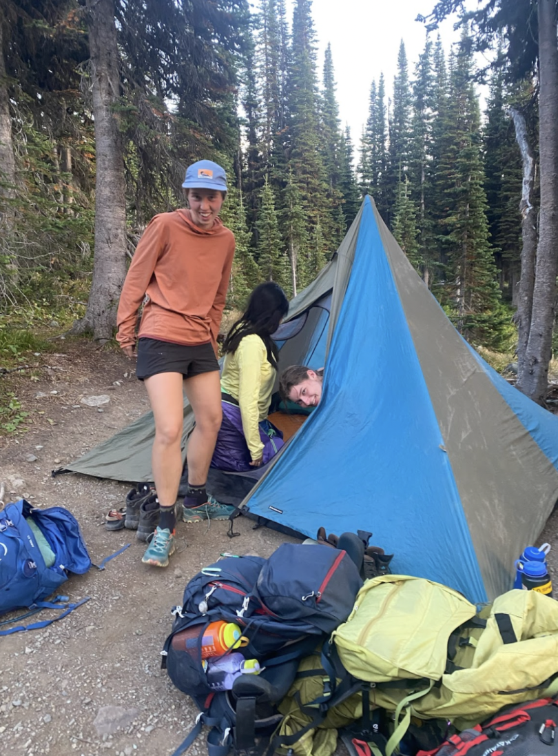 Two women sit inside a camping tent that looks like a circus tent, while another woman climbs out of the tent. Backpacks sit right outside of the tent. Two of the women are looking towards the camera, while the third is turned away.