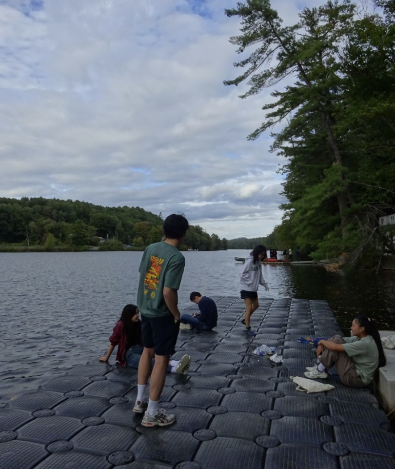 some college students are relaxing on a floating dock. There is a small hill and a river in the background. The sky is cloudy.