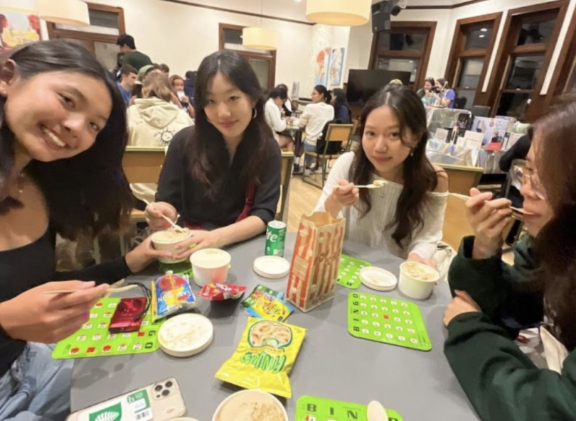 a group of four girls sit around a small table smiling at the camera. There are bingo cards and different snacks and drinks on the table. There are many people also sitting at tables in the background.