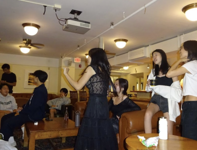 a group of college students gather in a dim karaoke room in a basement. Some are standing and dancing, others are sitting and singing. They appear to be having fun.