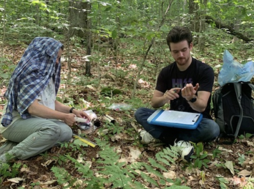 two people sitting next to each other in a forest. The woman is holding a piece of PVC pipe and looking at the man who is holding something in his hand. On his lap is a clipboard, and next to him is a backpack with a large, blue bag poking out the top.