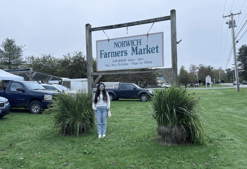 A girl with brown hair wearing a white long-sleeve top with blue jeans standing underneath a sign that reads "Norwich Farmers Market Saturday 9 am - 1 pm May thru October - Rain or Shine".