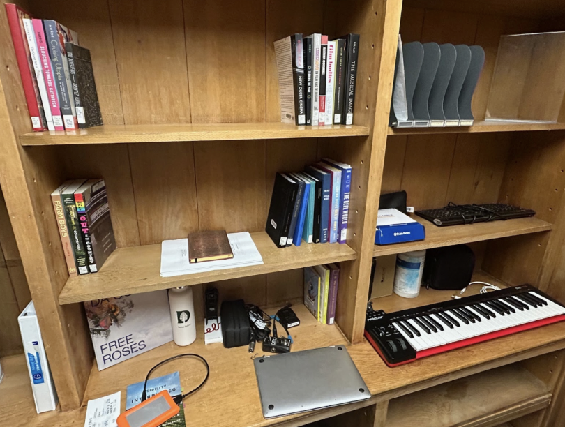 There are wooden bookshelves with books scattered across a few rows. In the bottom right, there is an electric piano with other electronic equipment.