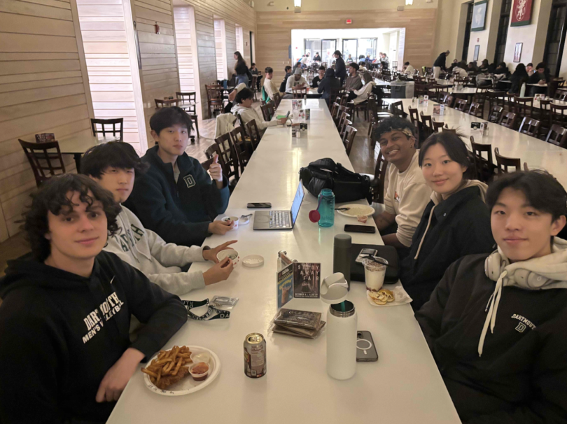6 college students sit facing each other at a table in a large dining hall. They have various fried foods in front of them. 