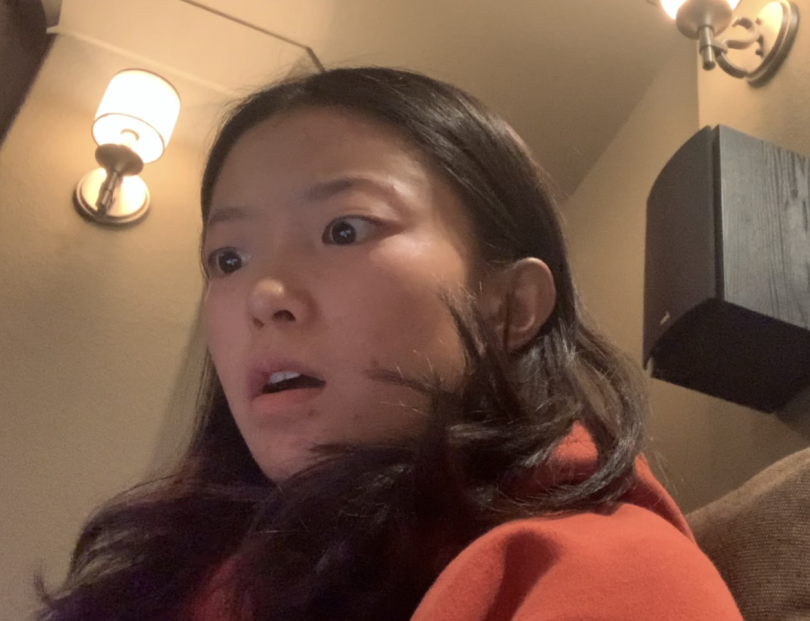 An image of me, Crystal, with a look of shock on my face. I am sitting in a chair in a room and there are wall fixture lights behind me. I am also wearing a red hoodie.