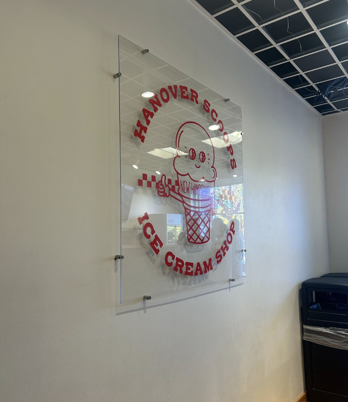 A glass decal inside of Hanover Scoops. It's a red print on glass that says "Hanover Scoops: Ice Cream Shop". In the middle is an ice cream cone with eyes that says New Hampshire and is holding a thumbs up.