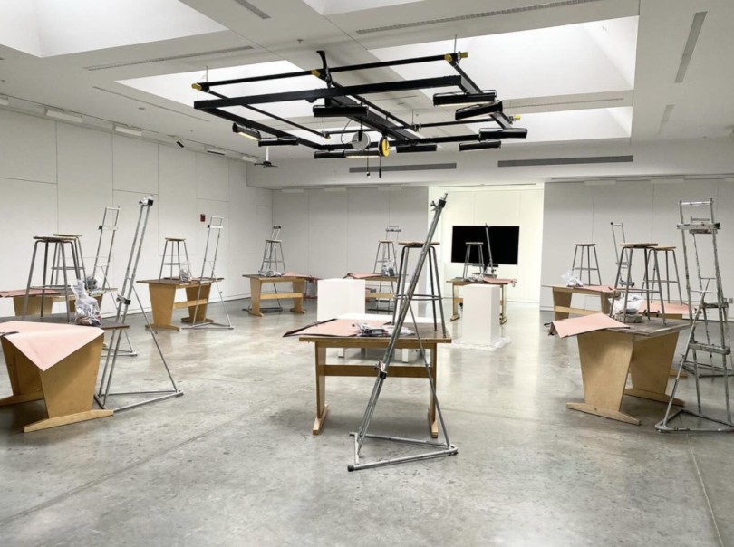 A brightly lit drawing studio with wooden desks and stools. 