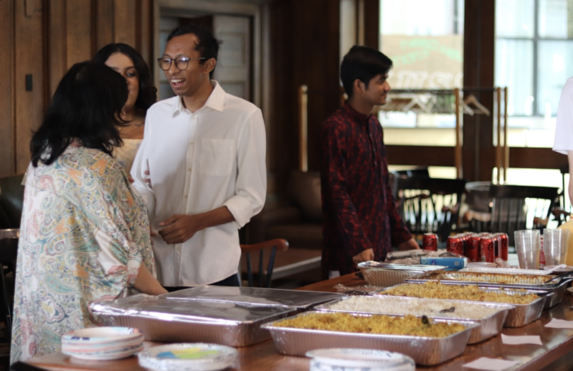 Picture portraying several students and members of the community wearing colorful Bangladeshi traditional outfits. In the picture, there's a table with several trays of food on top of it varying from rice, chicken, tofu, and more.