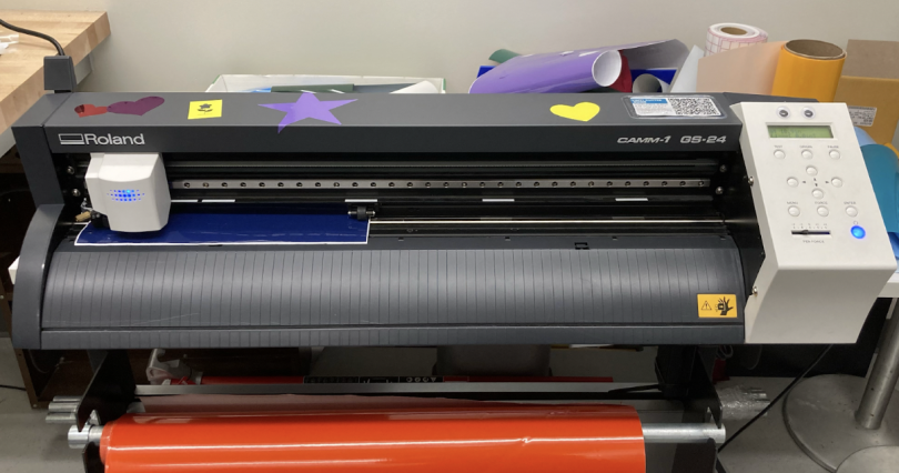 A vinyl cutting machine, the background contains a variety of colored vinyl rolls.