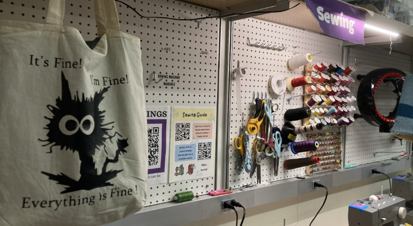 Sewing machines are on a table in the bottom right corner, the wall is covered with spools of thread, fabric shears, and flyers with sewing guides on them. In the foreground is a tote bag with a vinyl print of a cat with the text "It's Fine! I'm Fine! Eve