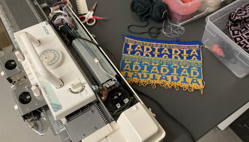 A flatbed knitting machine is in the bottom left corner. A knitting sample of dark blue, light blue, and yellow stitches spell out "ADIADIAD" in a repeating pattern separated by stripes.