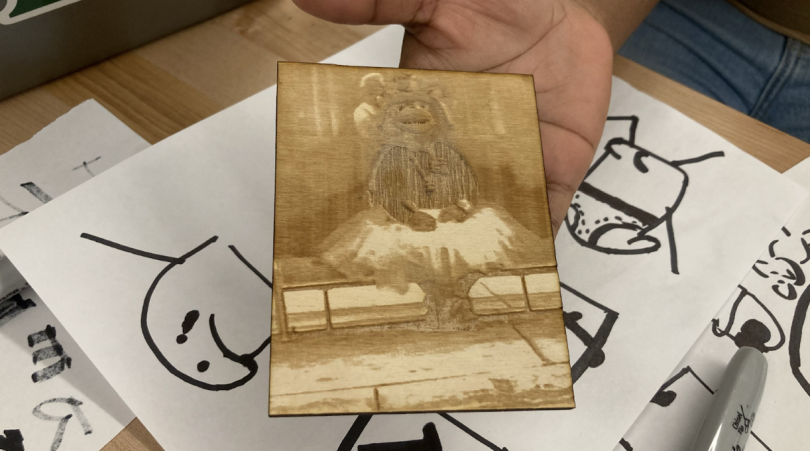 A rectangular piece of wood with a bear in a tutu etched into its surface. The background is a table covered with sheets of paper and sharpie drawings.