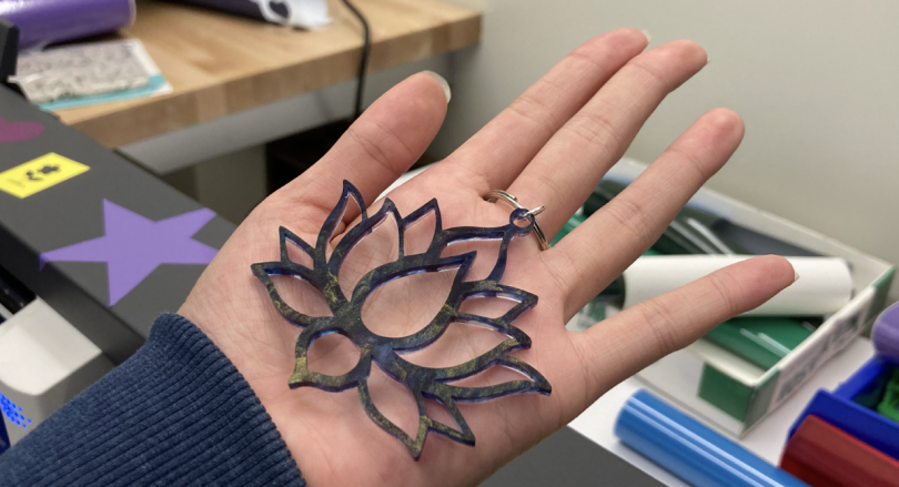 A lotus keychain cut out of blue and gold colored acrylic held in the palm of Tina's hand. In the background is the vinyl cutter and rolls of green, blue, and purple colored vinyl.