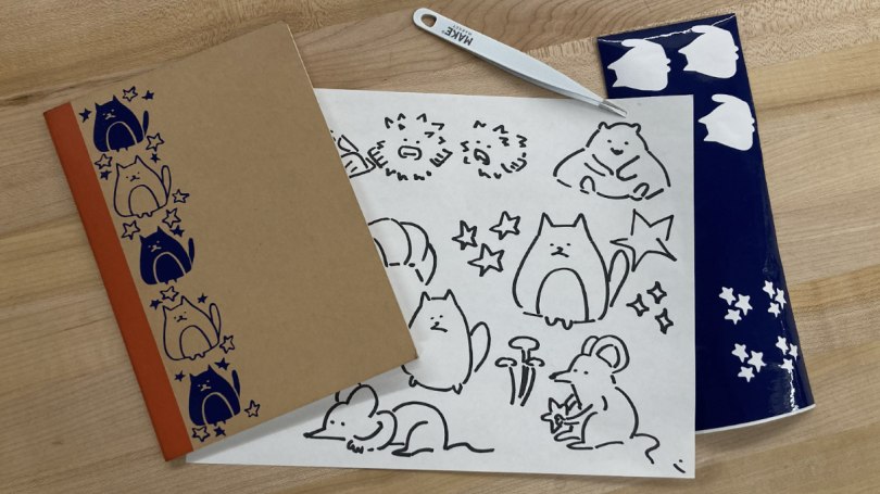 Left is a brown notebook with an orange stripe and blue cat and star stickers along the left edge. Middle is the sheet of paper with my sticker ideas drawn in Sharpie. Left is the sheet of navy blue vinyl that my stickers were cut out of and a pair of twe