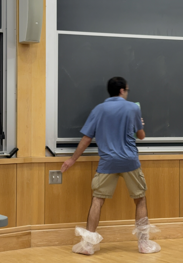 A man wearing plastic bags for shows with a blue sweater and khaki shorts writing on a board.
