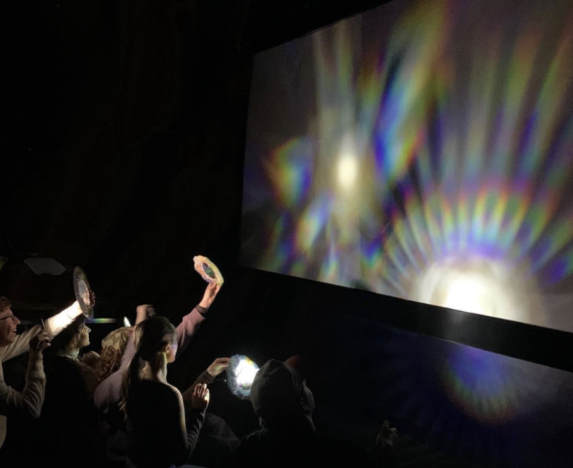 Students sit in the bottom right with flashlights and translucent paper. They shine light through the paper onto a white board which makes patterns of rainbows on the screen.