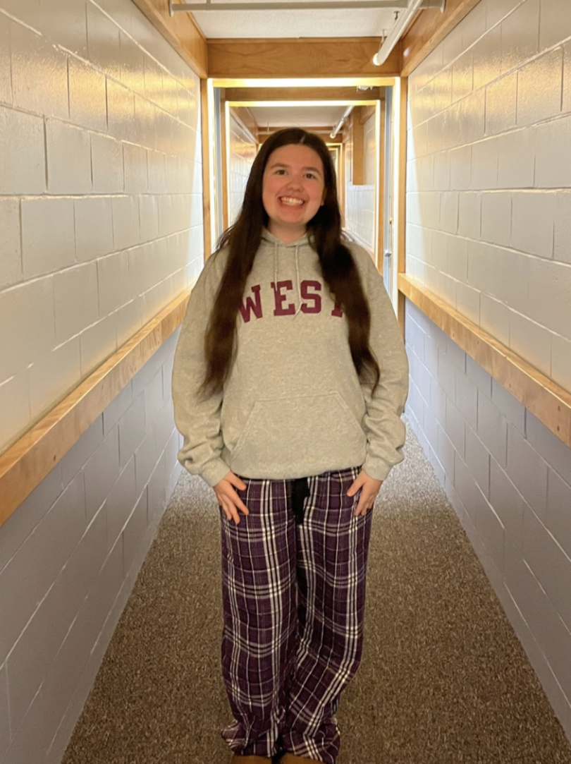 A girl with brown hair wearing purple plaid pants and a gray sweatshirt that says "West" standing in a hallway.
