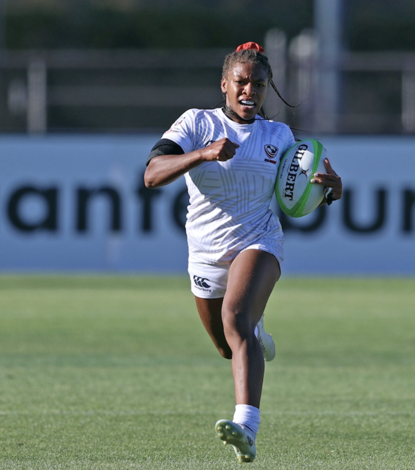 A woman runs on a green field holding a rugby ball.
