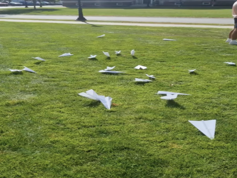 A picture portraying several paper airplanes laying in a green field. In the background some people can be seen as well as a street and a street lamp.