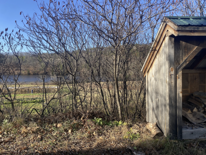 Half of an open storage hut with firewood stacked inside. Small trees and the river with mountains in the background.