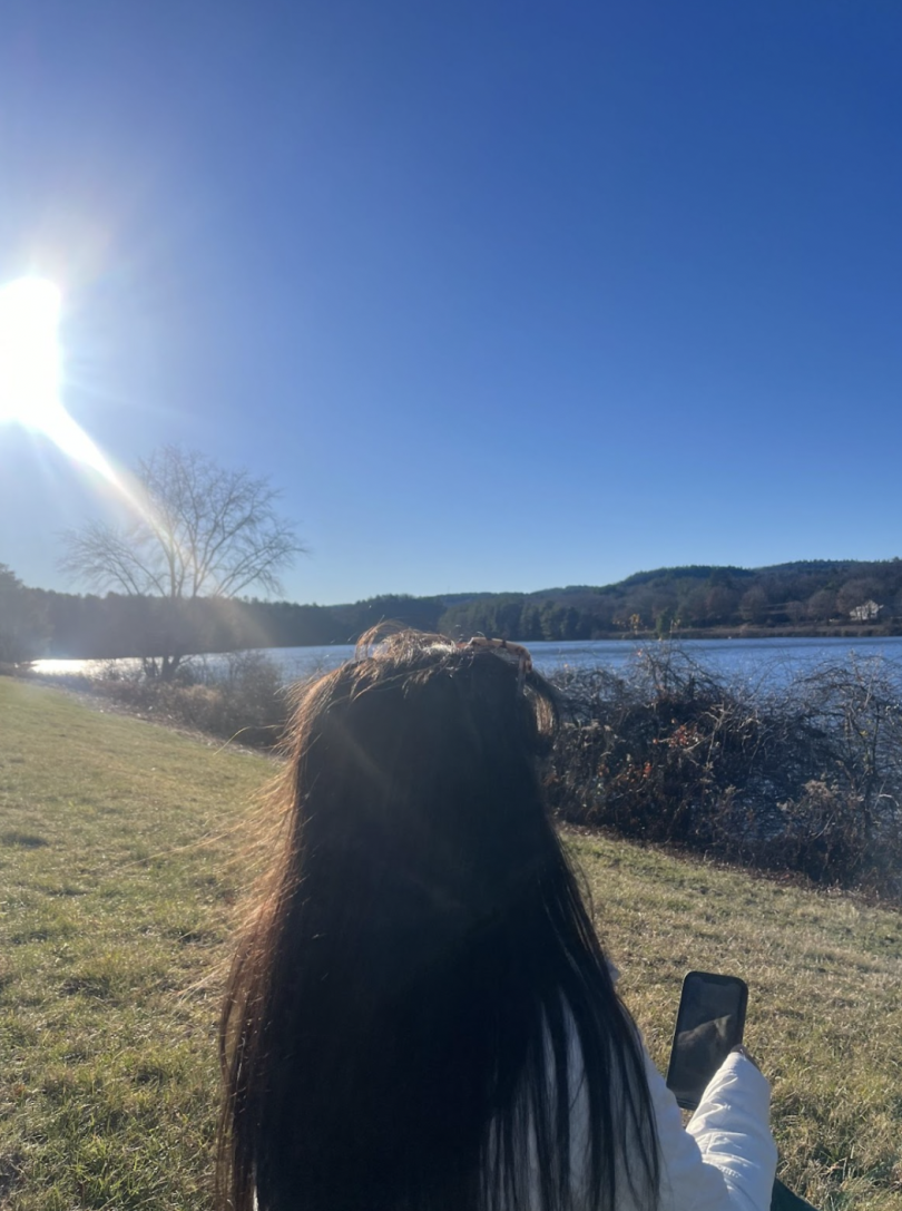 The back of a girl's head in front of a river. She is sitting on the grassy riverbank, with the sun shining.
