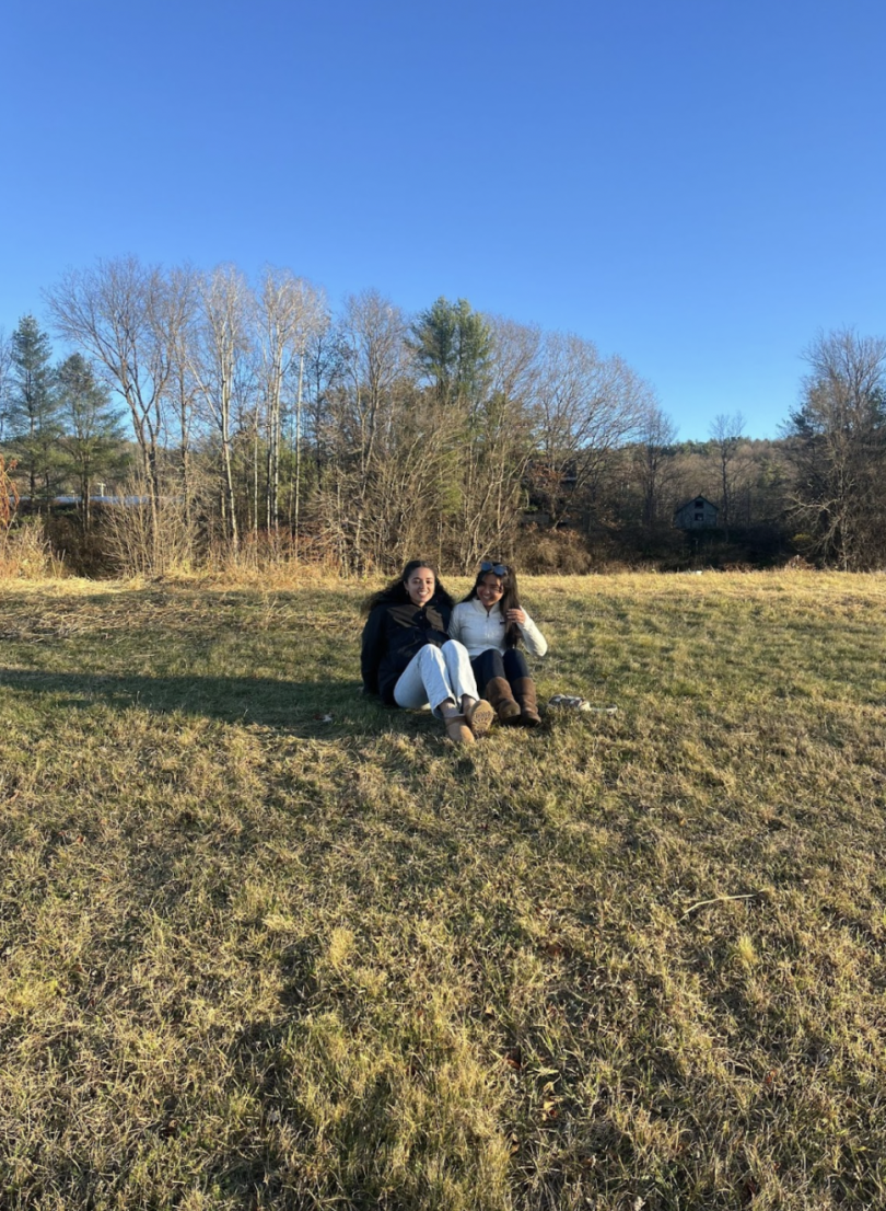 Two girls sit on a grassy field with trees and the blue sky in the background.