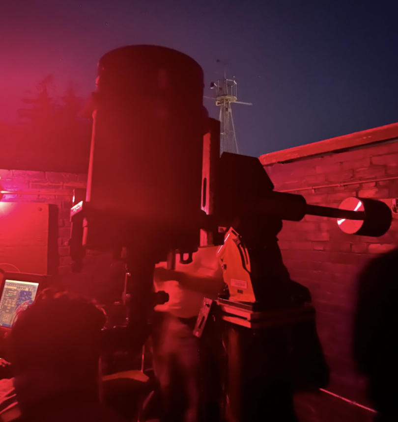 A large black telescope is trained toward the dark night sky and has a red hue.